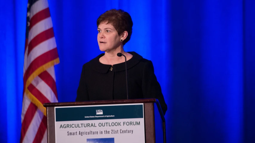 a woman standing at a podium with an american flag behind her