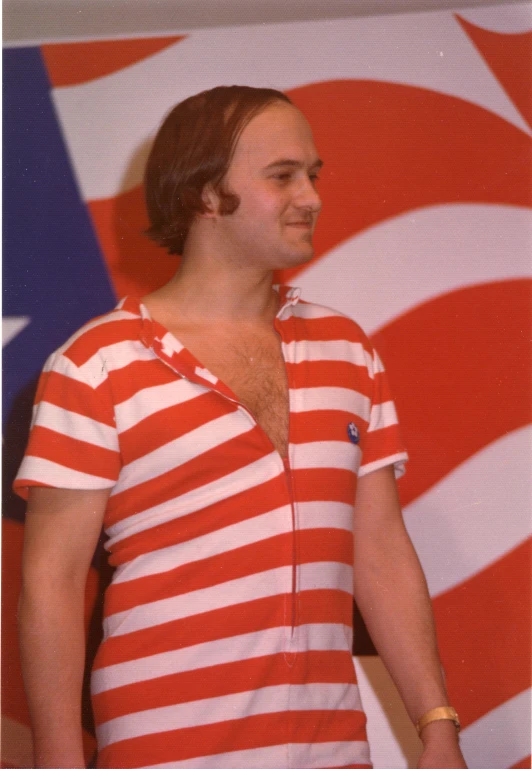 man in red and white striped shirt standing next to large american flag
