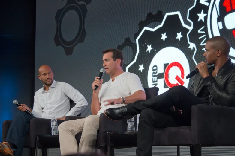 three men sitting on chairs while one holds a microphone in his hand