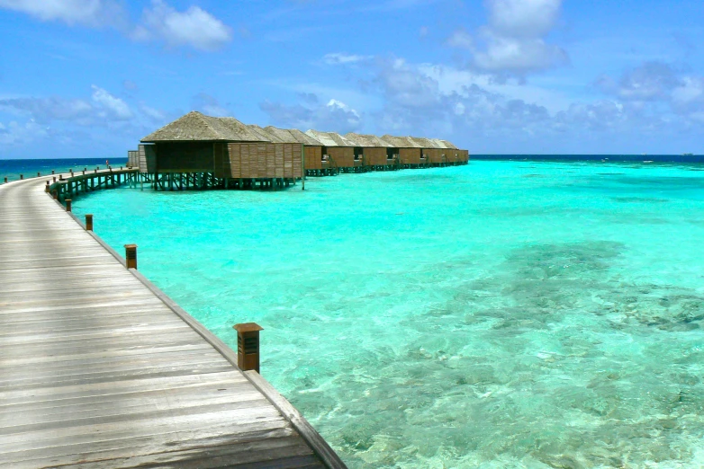 a boardwalk extends into the turquoise ocean towards water with huts in the background