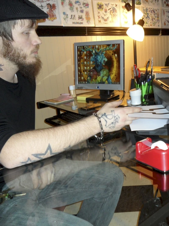 a tattooed man sitting at his desk pointing at a computer