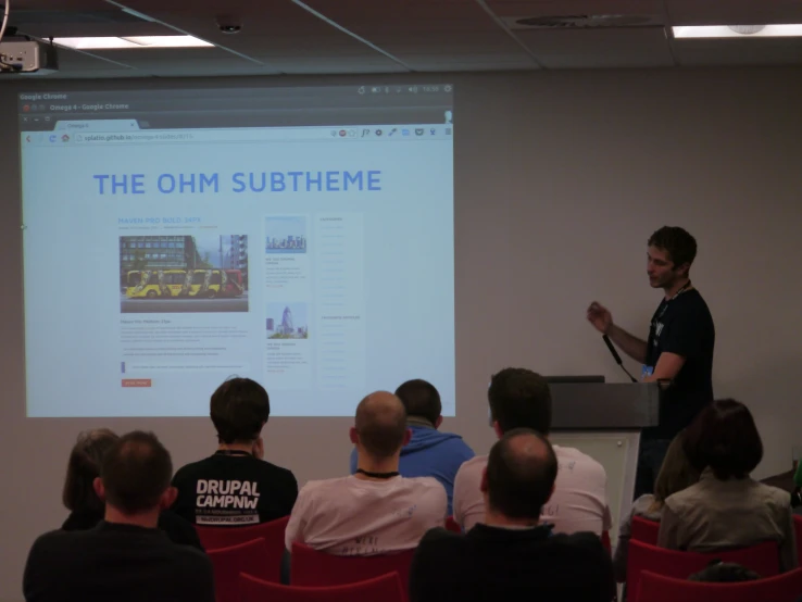 a man stands at the front of an auditorium with his microphone in front of a screen giving a lecture