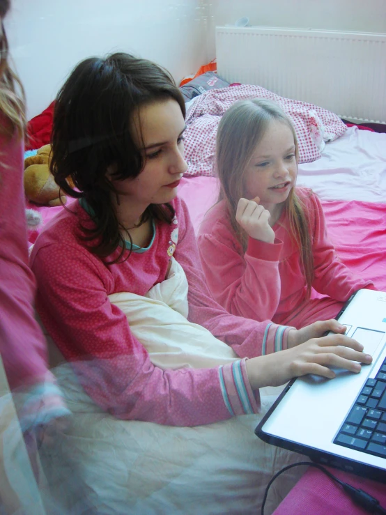 two little girls are sitting on their bed looking at a laptop