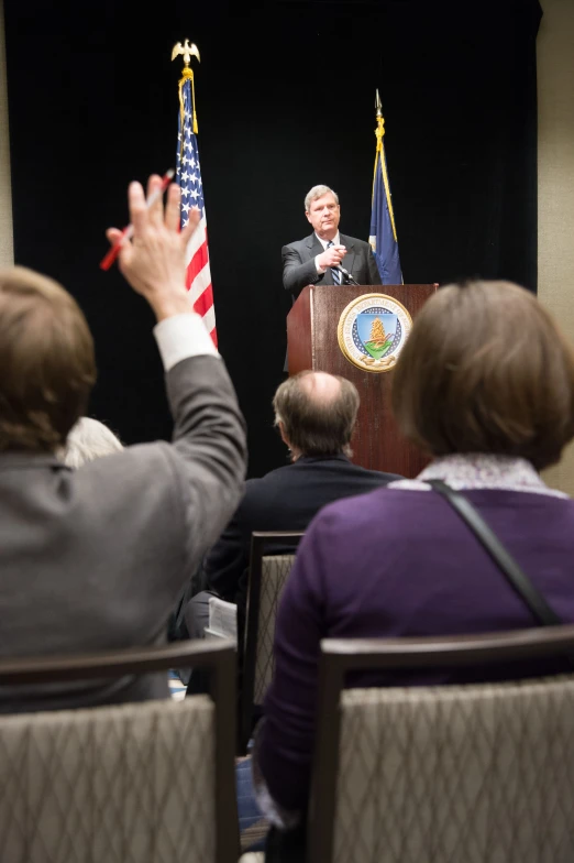 a man in suit sitting in front of a microphone with his hands up to speak