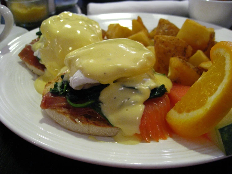 an egg benedict sandwich with tomatoes, orange wedges and potatoes on a white plate