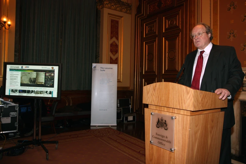 a man in a business suit standing at a podium in front of a tv