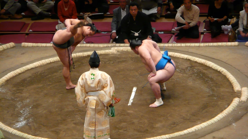 two sumo wrestlers standing in an arena while one man looks on