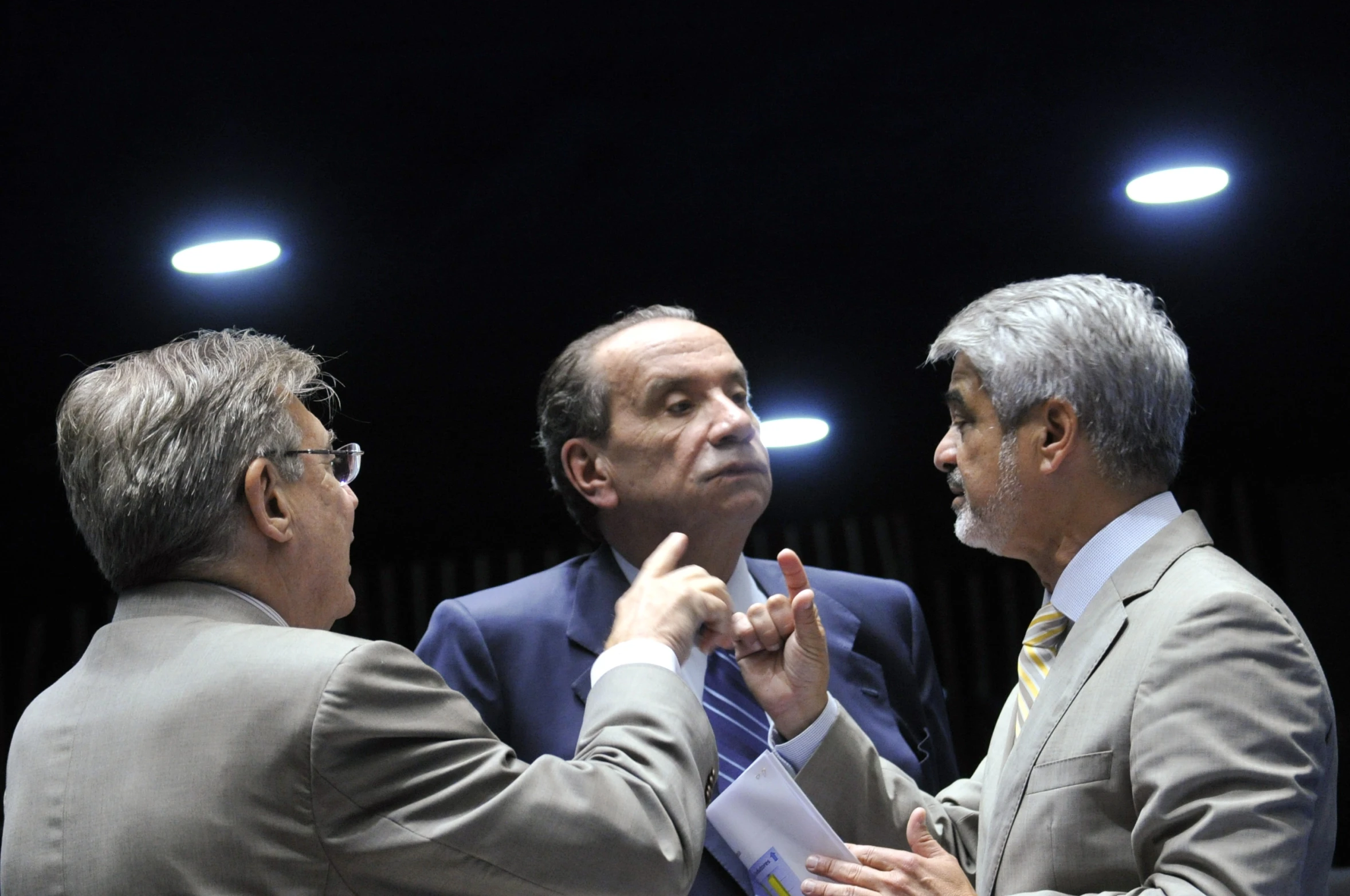 three people wearing suits talk together as one man watches from a stage