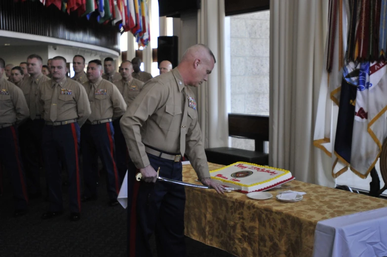 a military man cuts a cake on a table with other soldiers behind him