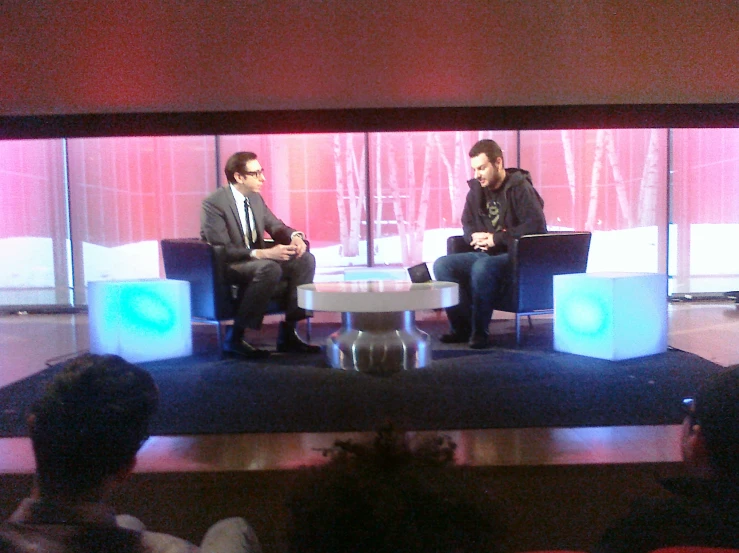 two men sit in chairs with a television and are on stage