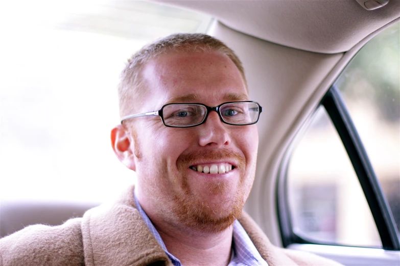 a smiling man with glasses sits in a car