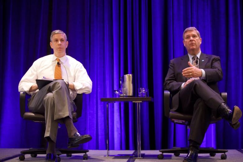 two men sit on stage with chairs in front of them