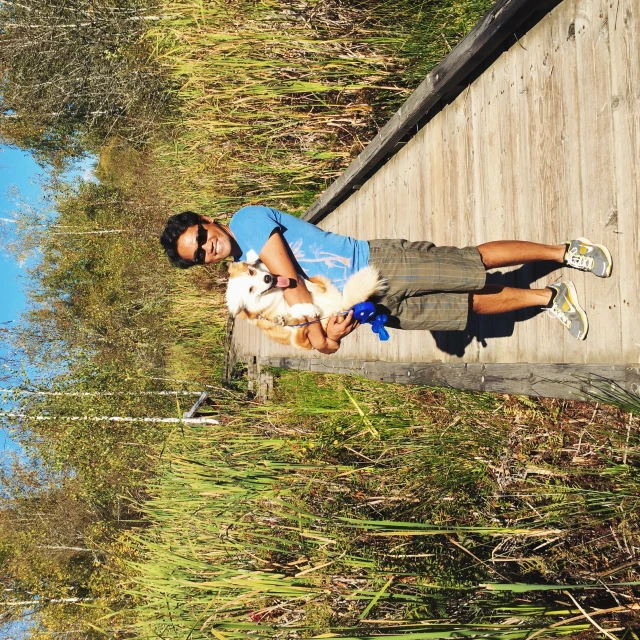 a young man holding a dog on a boardwalk next to tall grass