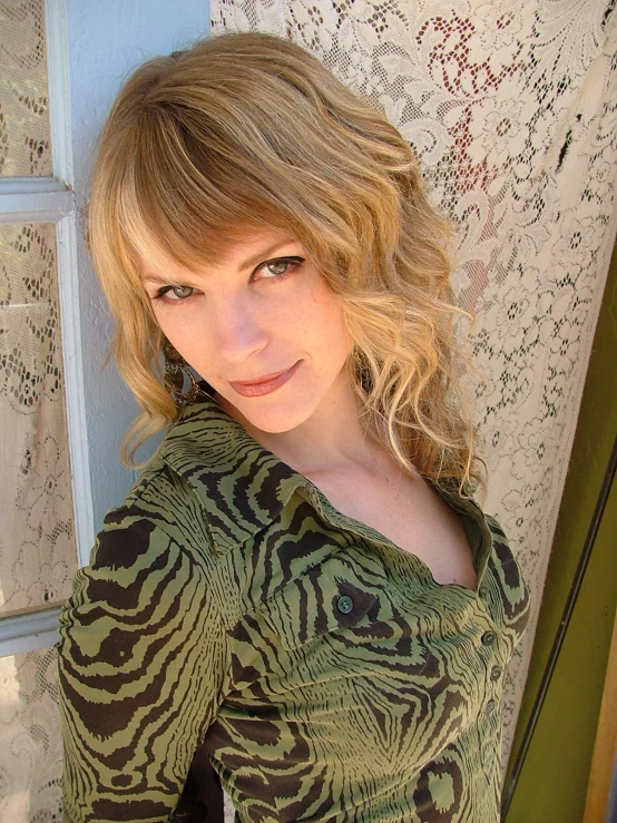 young blonde woman in an allover green and brown top posing by a window