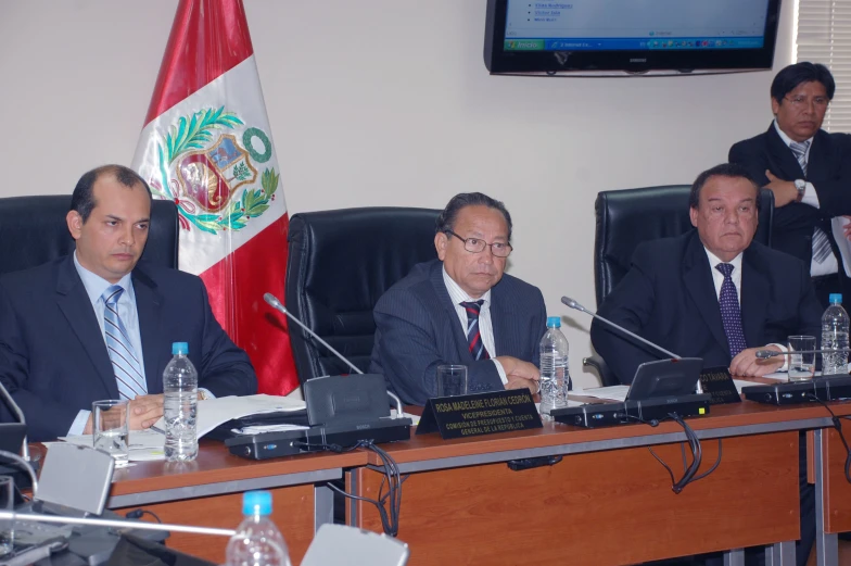 three men in business suits sit at desks with microphones