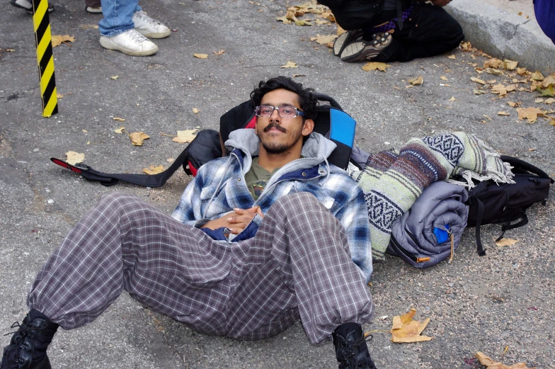 a man with glasses sits on the ground among backpacks