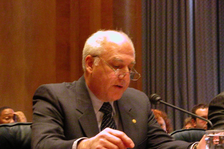 an older man in a suit talks in front of microphones