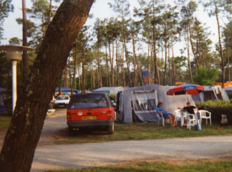 two tents with people seated outside near a tree and parked car