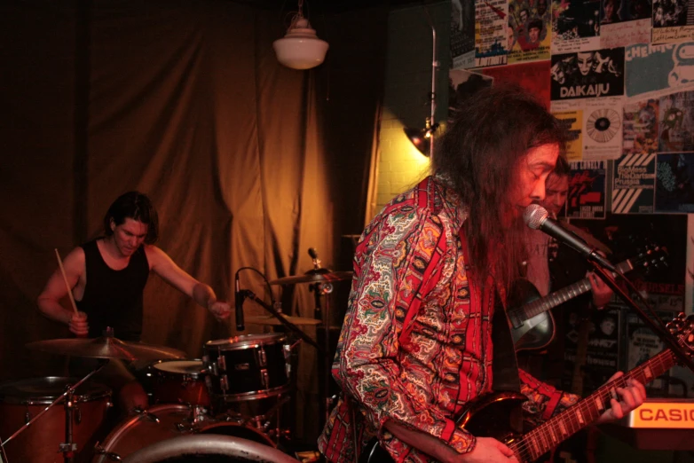 a man with long hair is singing into a microphone while another man plays the drums in front of him