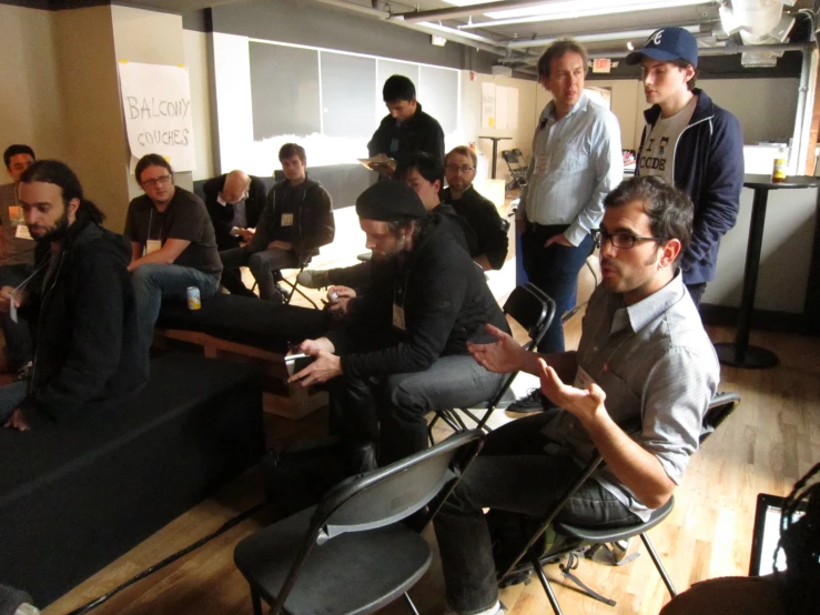 some men sitting at tables in an office with many other people standing