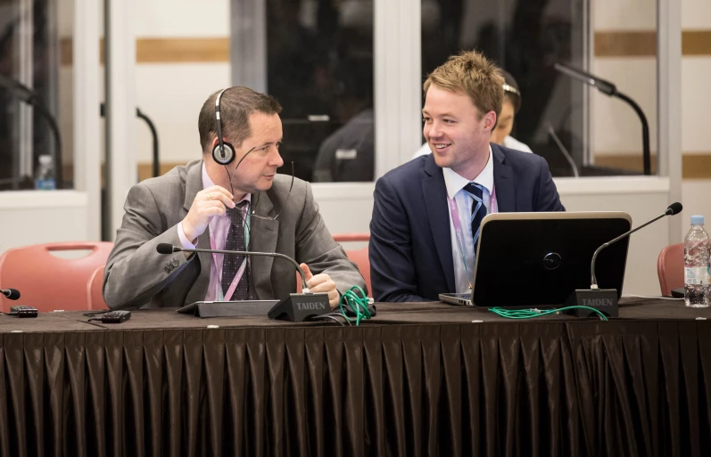 two men sit at a table looking off into the distance, with their hands on their chins