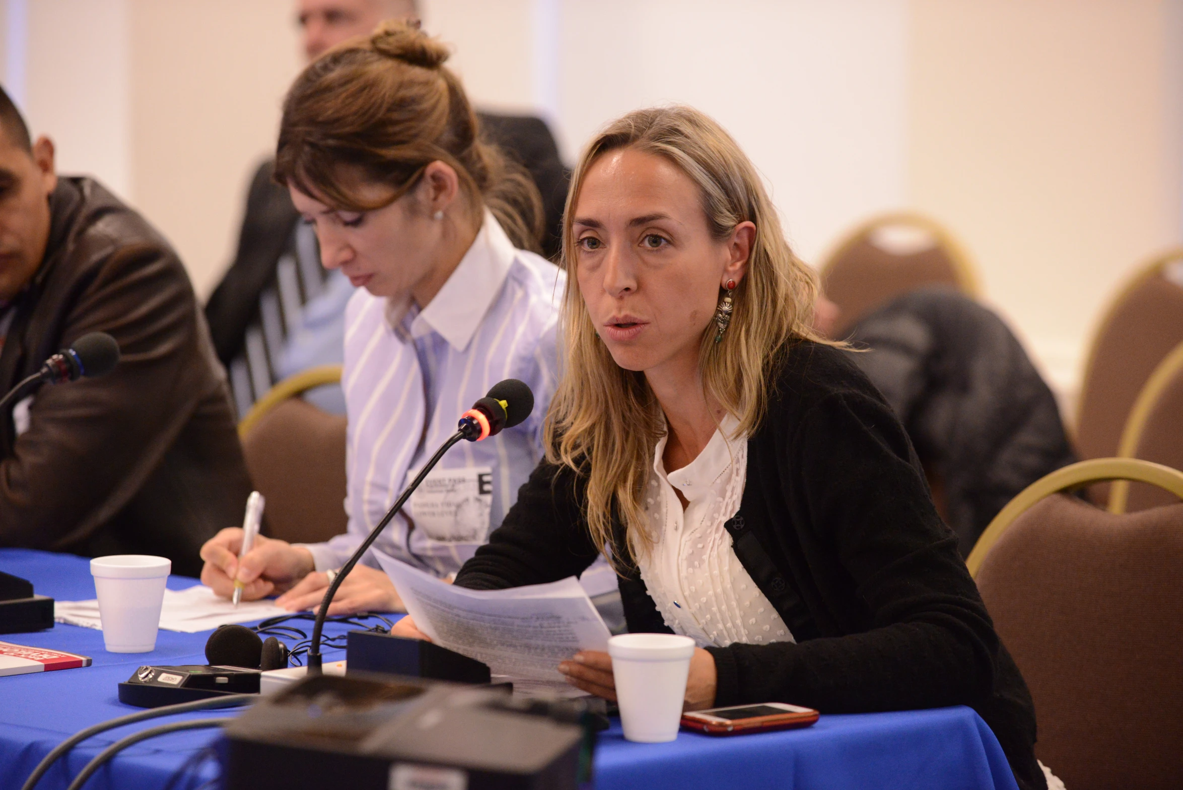 a woman sits at a blue table with two microphones