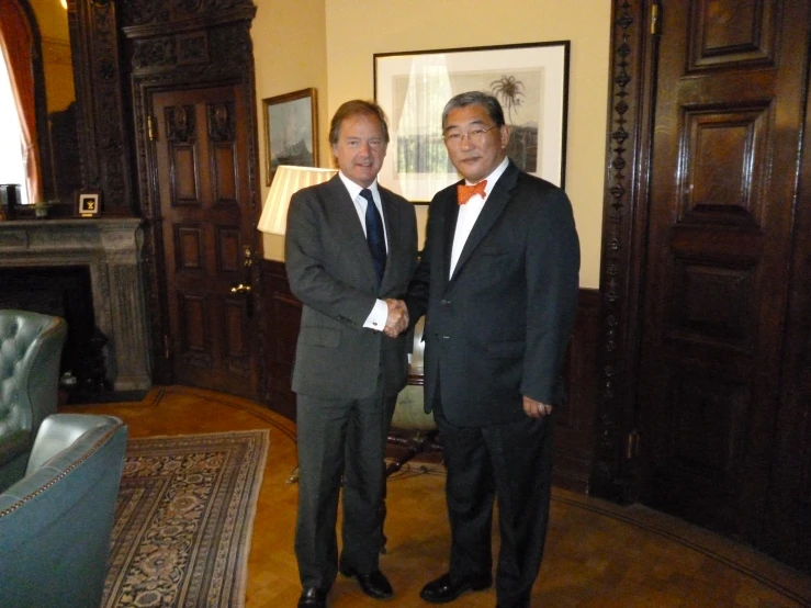 two men in suits and ties are standing in a room with wood floors and an ornate framed fireplace