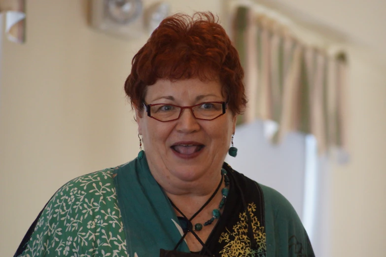 a red - haired woman in glasses smiles, she wears green, and a green flowered shirt