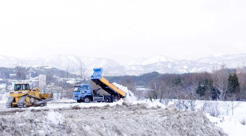 a truck and dump truck with large tires are dumping sand from a snowy hill