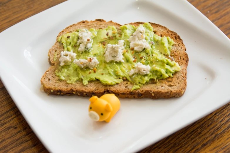 a square plate holds a toast with guacamole, cheese, and an orange