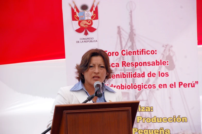 an older woman speaking at a podium with red flag behind her