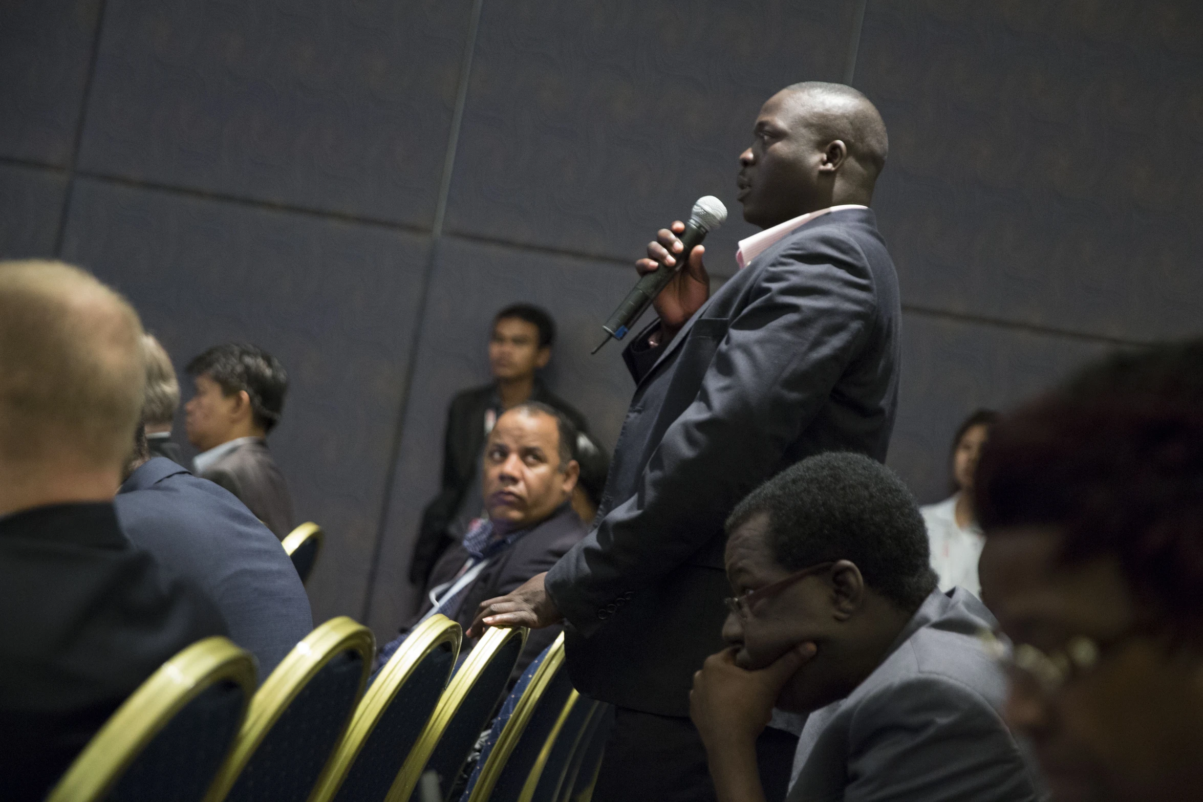 man standing on stage with microphone in foreground while others wait for him