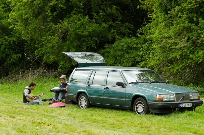 two people sit on the grass near their car with their luggage