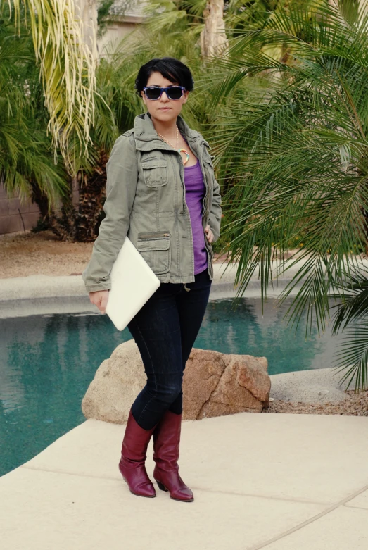 a woman wearing red boots, black jeans and a green jacket is standing near a pool with a book in her hands