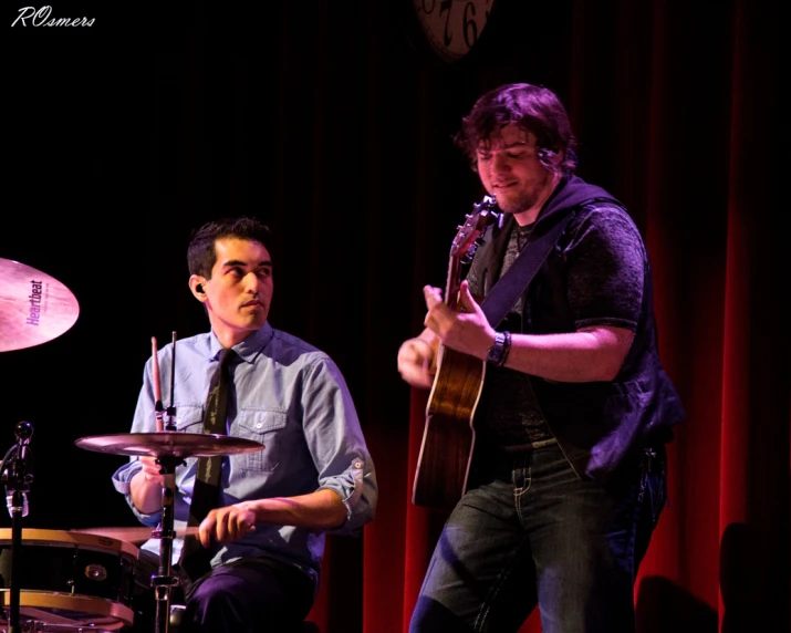 a man playing guitar with a man sitting beside him on the drum set