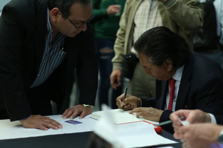 two men are sitting at a table while one signs a paper