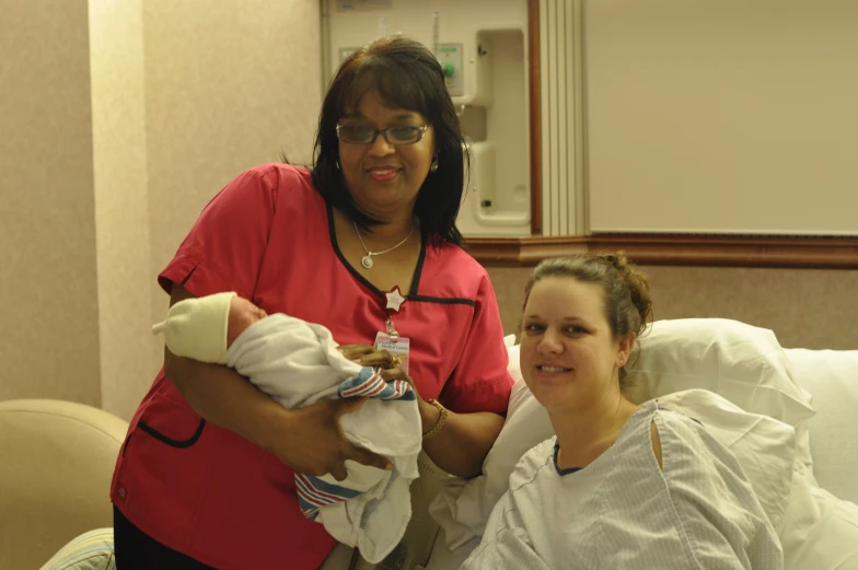 two women smile while holding a baby in a hospital bed