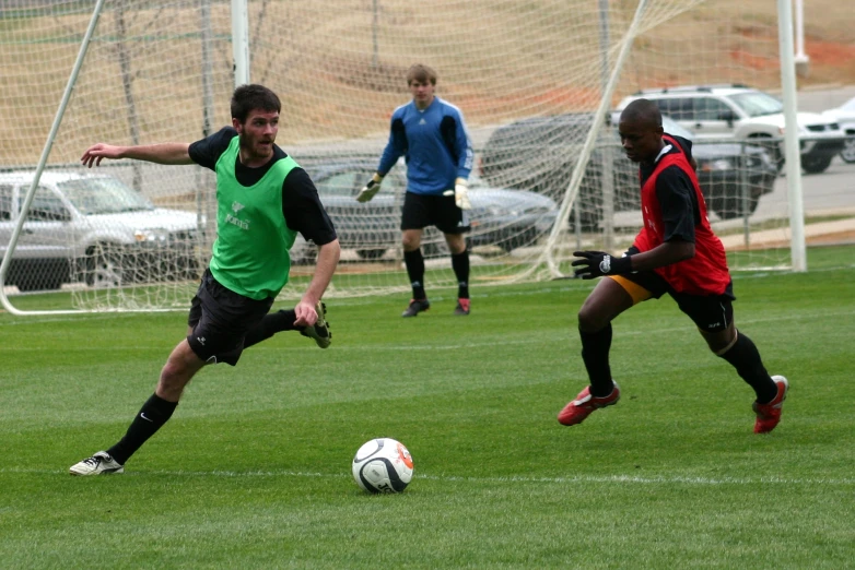 three men playing soccer on green grass with cars behind them