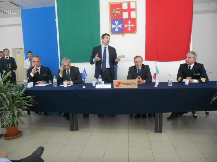 men wearing suits sitting at a table with flags on the wall behind them