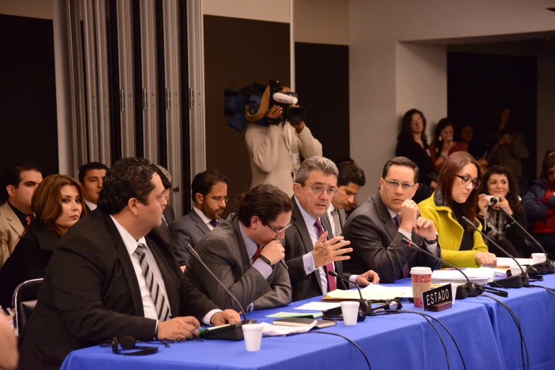 several people sitting at a blue table with microphones in front of them