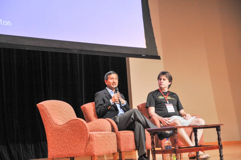 two men sitting down on chairs speaking on a projector screen