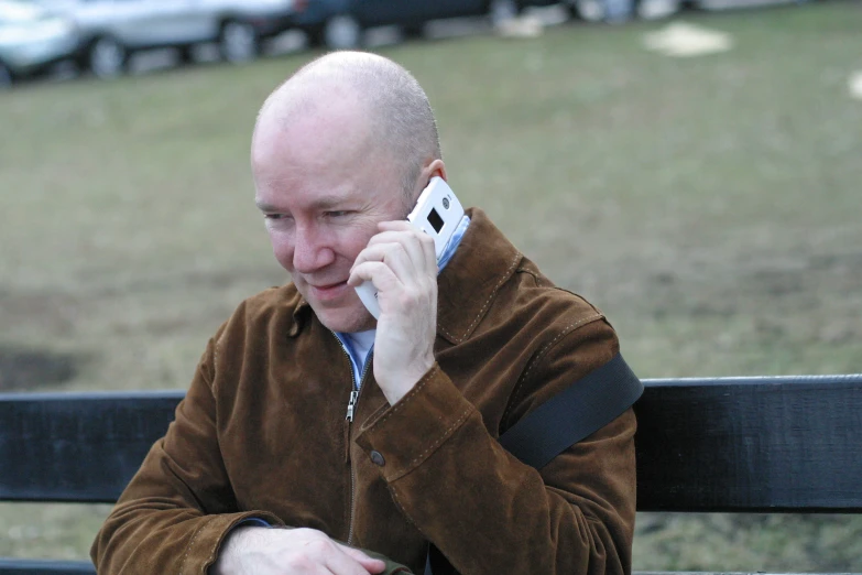 a bald man sitting on a park bench talking on his cell phone