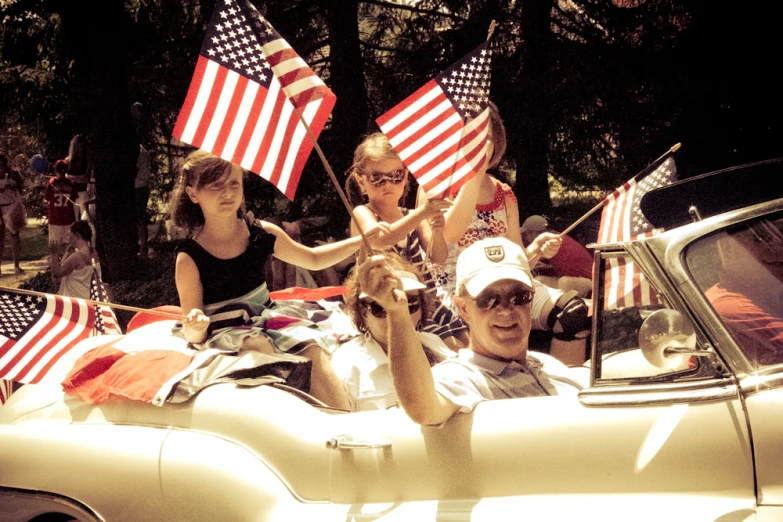 people sitting in an open topped car while waving american flags