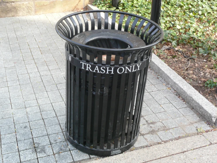 a trash can sitting on top of a sidewalk next to grass and bushes