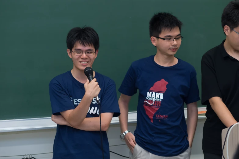 three young men are talking and holding microphones in front of a chalk board