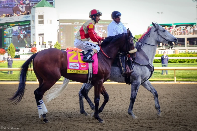 two men ride horses around a track in the sun