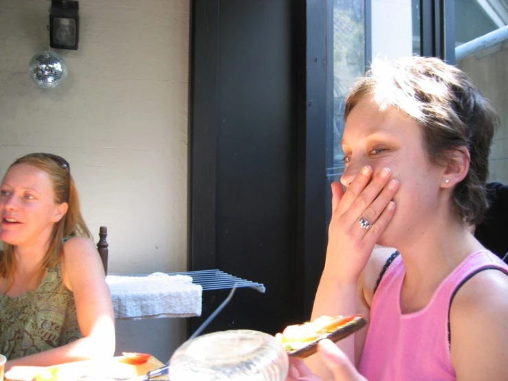 a lady covering her face outside while someone eats at the table