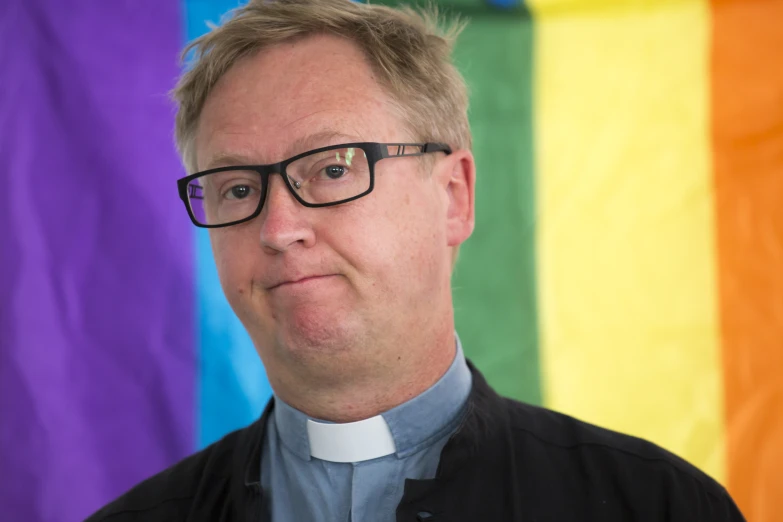 a man with glasses and a church uniform stands in front of a rainbow flag