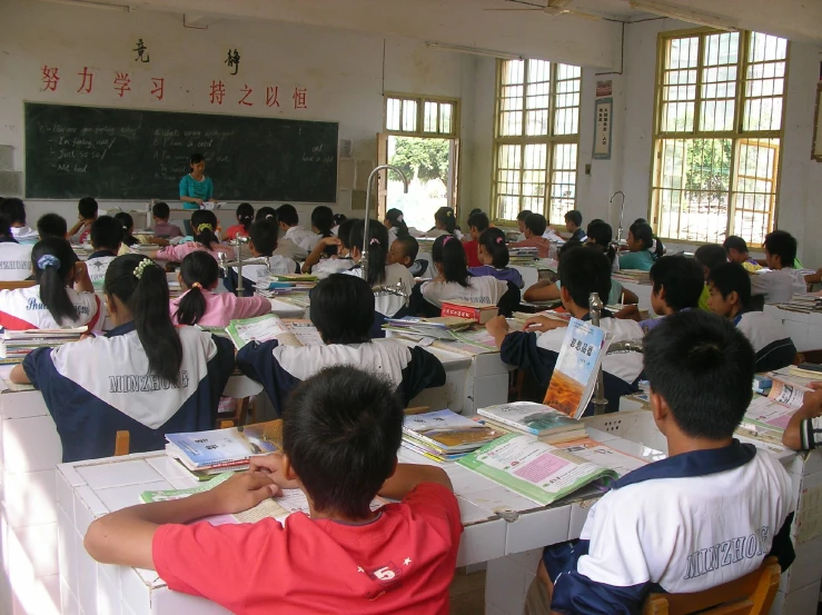 a school classroom with students sitting at desks and one teacher standing in front of the classroom writing on the blackboard