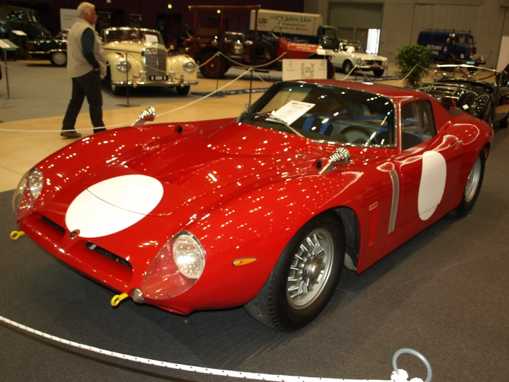 a vintage red sports car on display with an older man walking near it